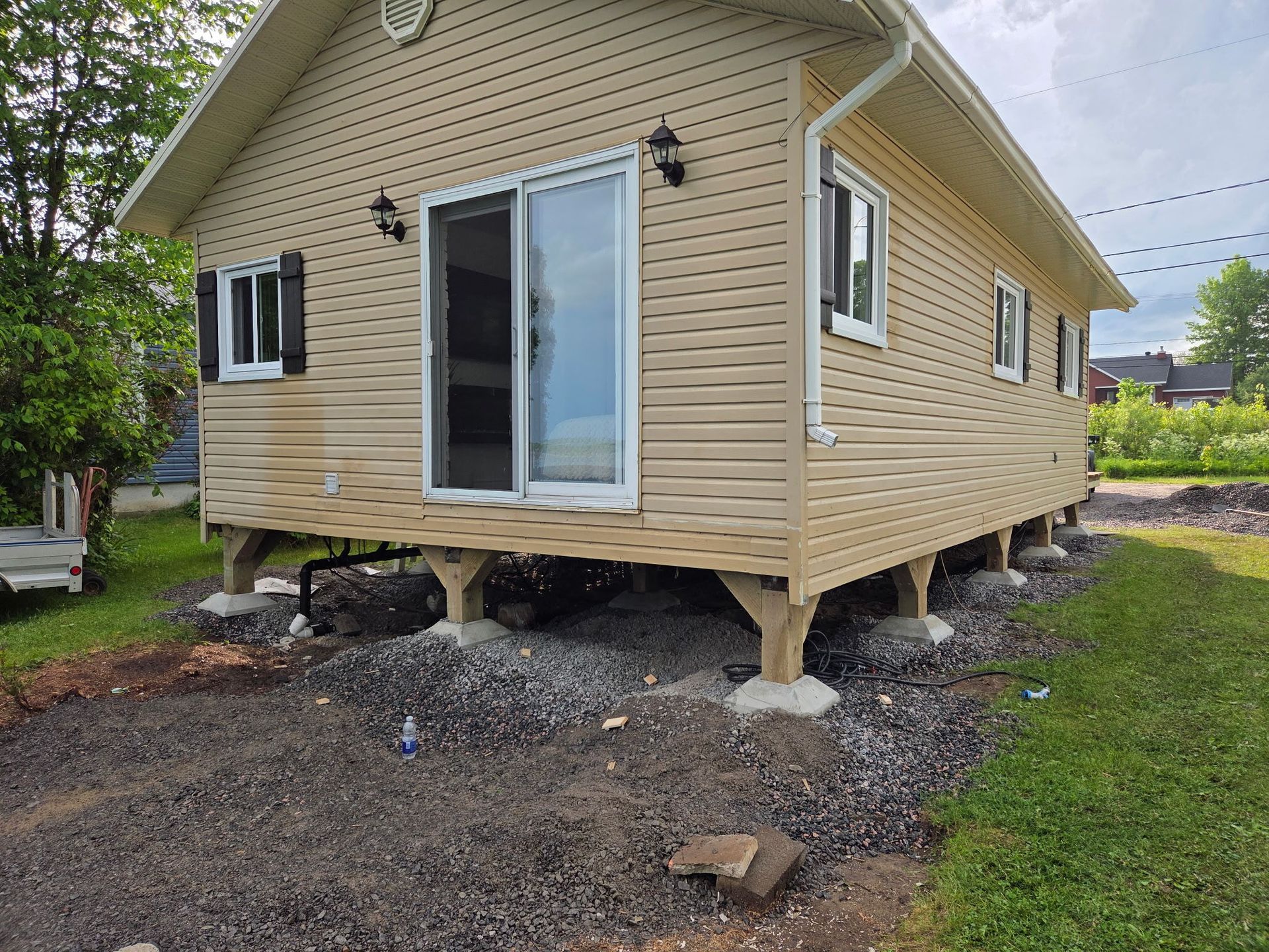 Maison beige sur pilotis au-dessus d'une allée de gravier, avec une porte coulissante en verre et de petites fenêtres.