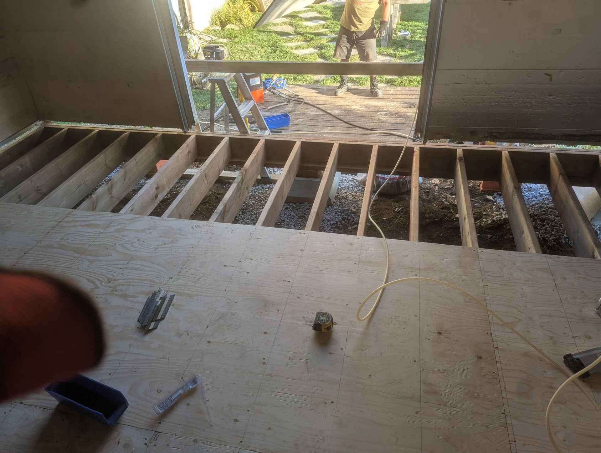 Pièce intérieure en travaux, solives apparentes. Sous-plancher en contreplaqué installé, porte ouverte sur l'extérieur.