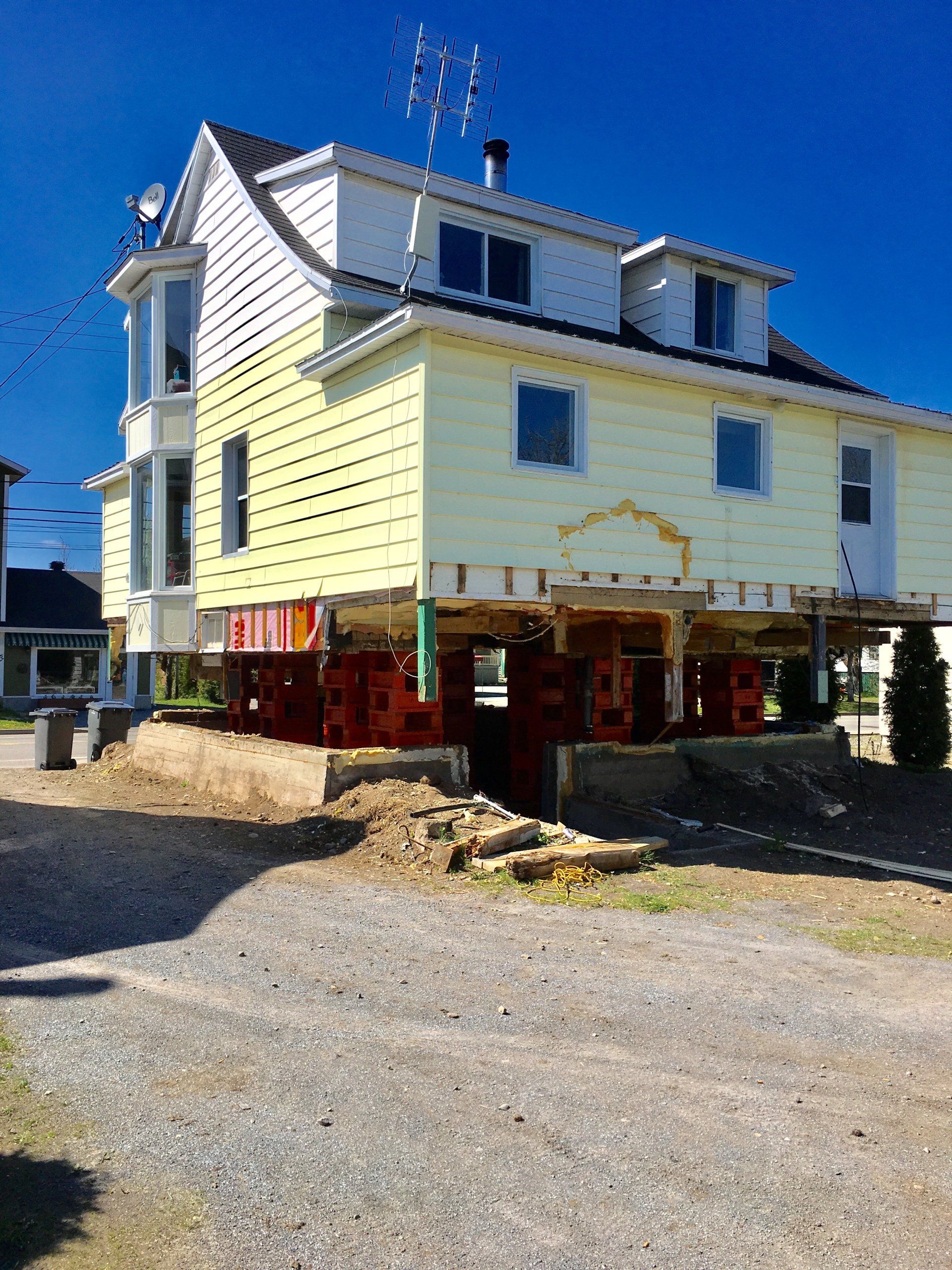 Maison surélevée sur pilotis ; bardage jaune, toit sombre, chantier.