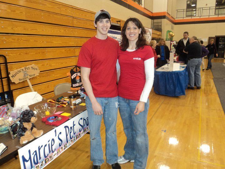 Two people stand in front of a pet shop table. They wear red shirts and jeans. Indoors, hardwood floor.
