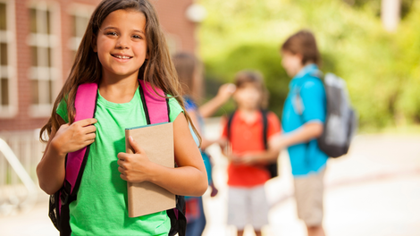 A young girl with a backpack is holding a book in front of a school building.