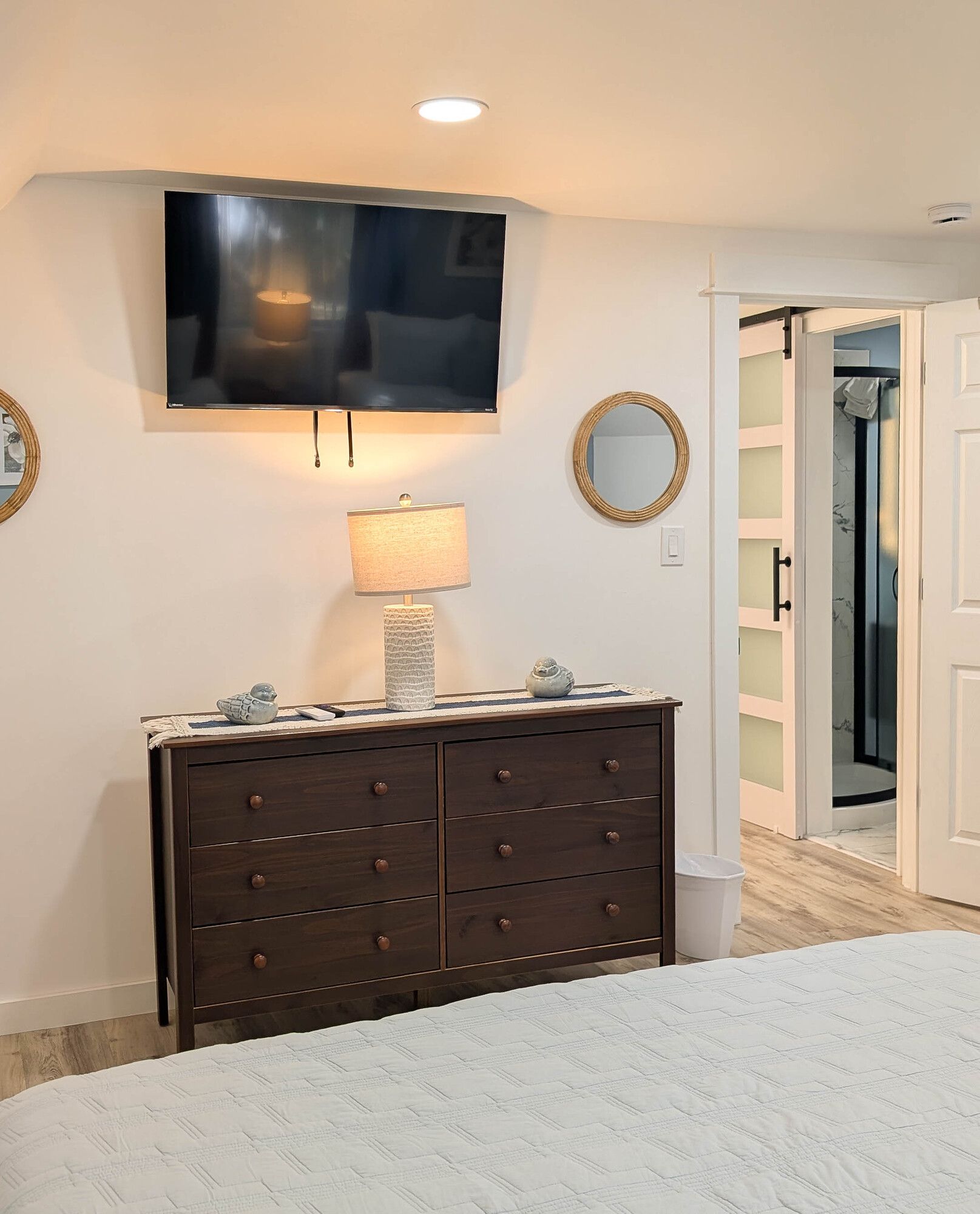 Bedroom with dresser, mounted TV, round mirror, and open doorway to bathroom.