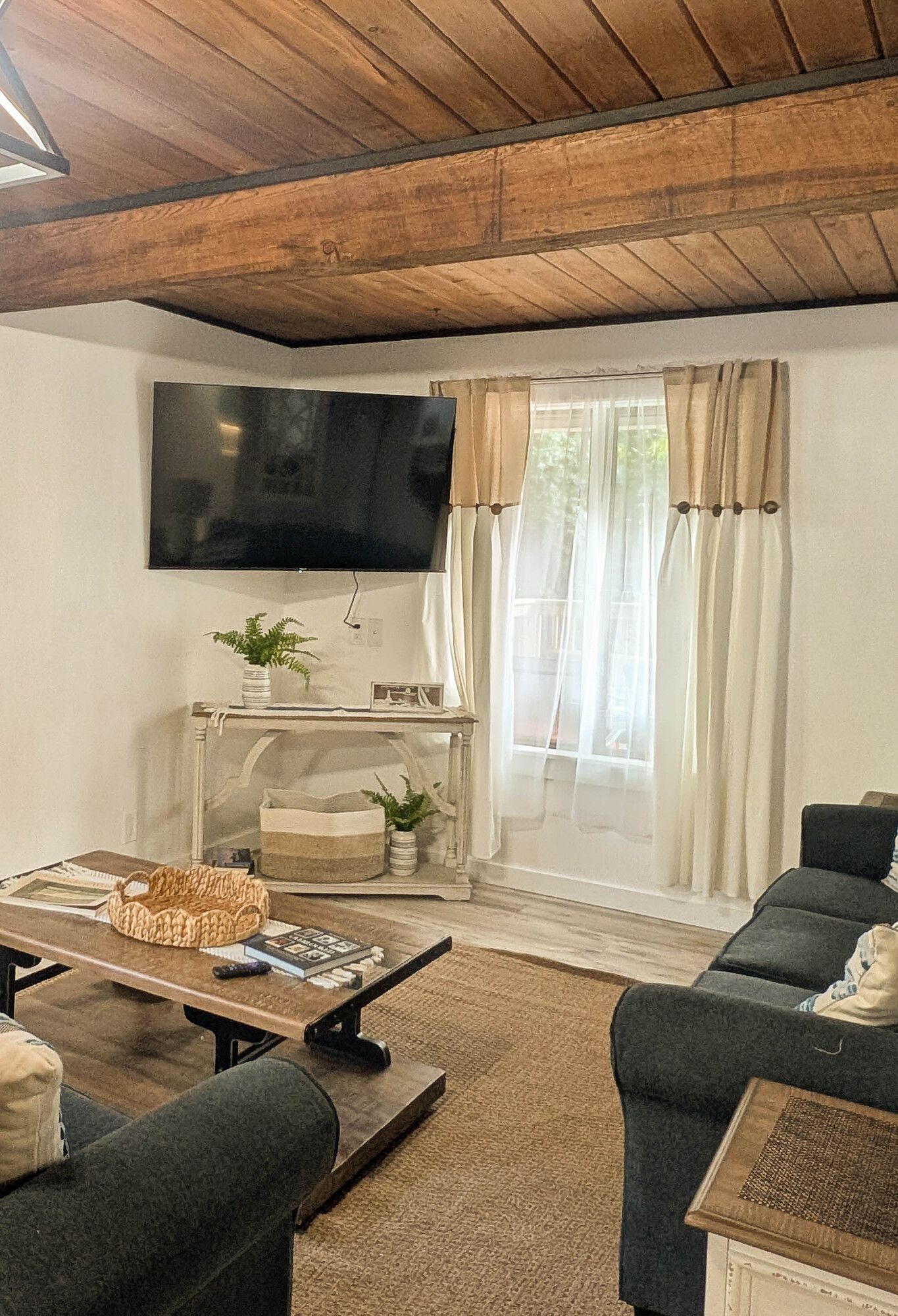 Living room with a wooden ceiling, TV, sofa, and a window with beige and white curtains.