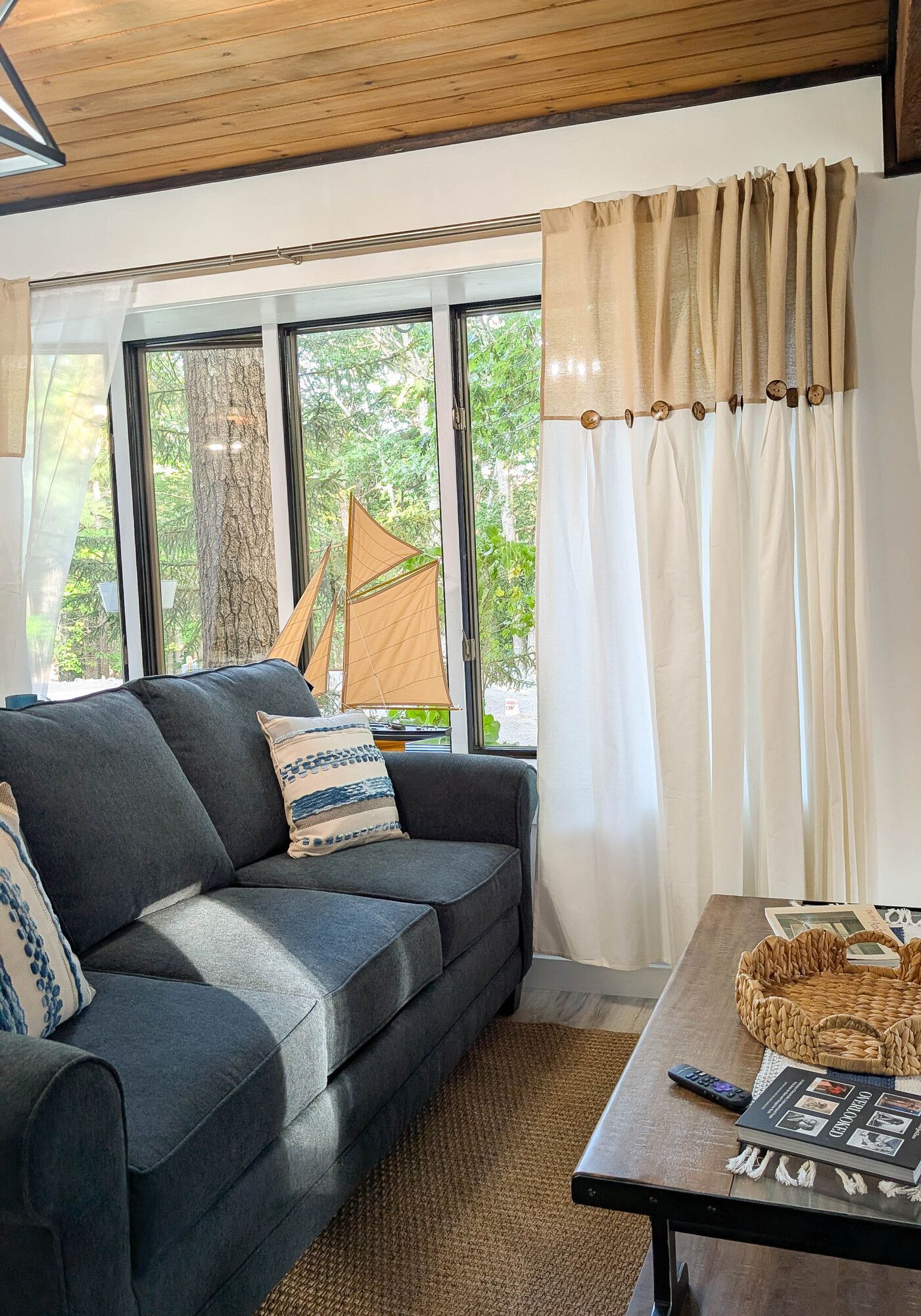 Cozy cabin interior with a blue couch, burlap and white curtains, and a window overlooking trees.