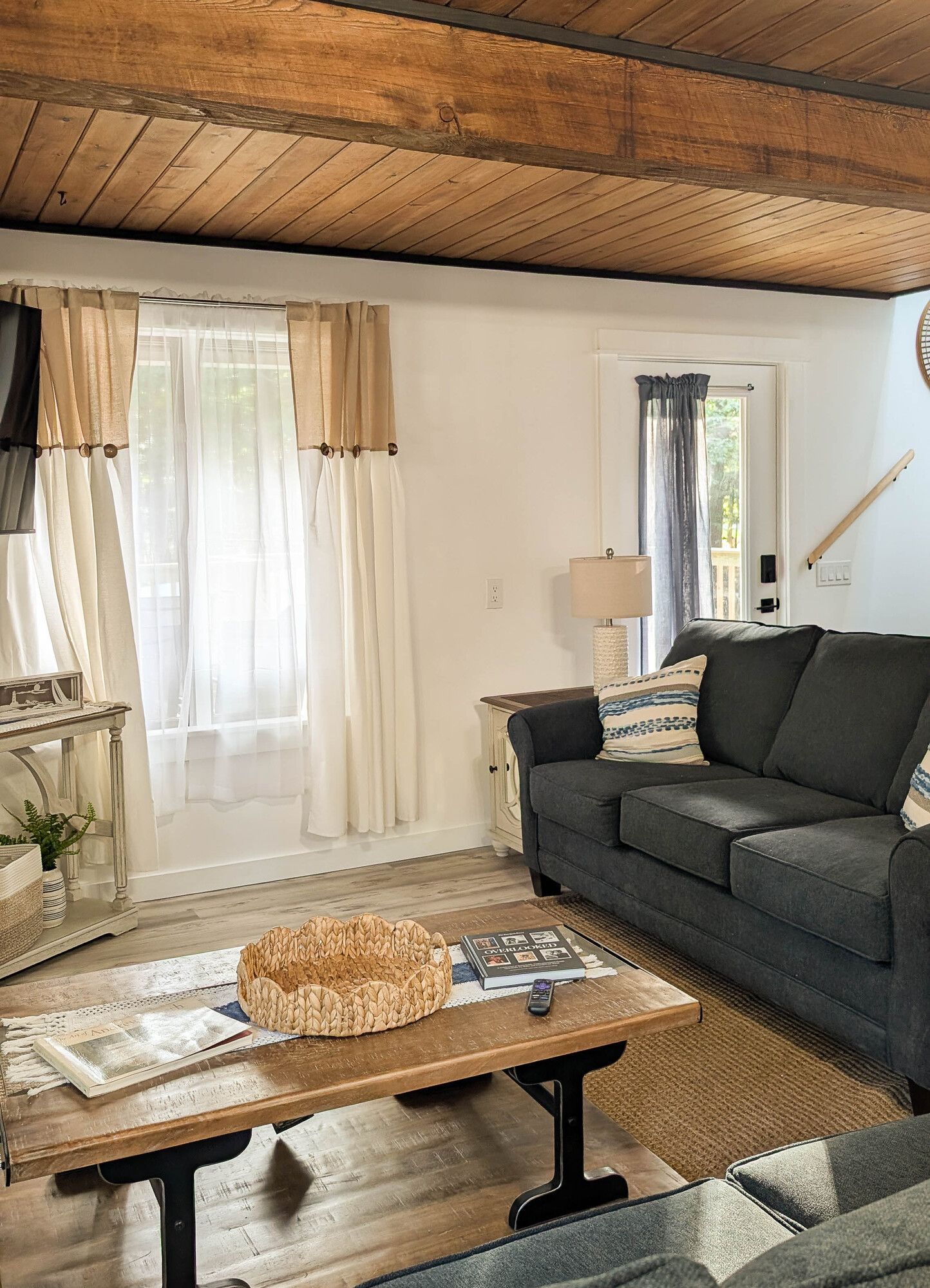 Cozy living room with a blue sofa, wooden ceiling beams, and a window with sheer curtains.