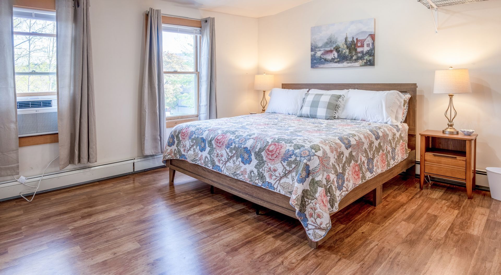 A bedroom with a king size bed , two nightstands , two windows and hardwood floors.