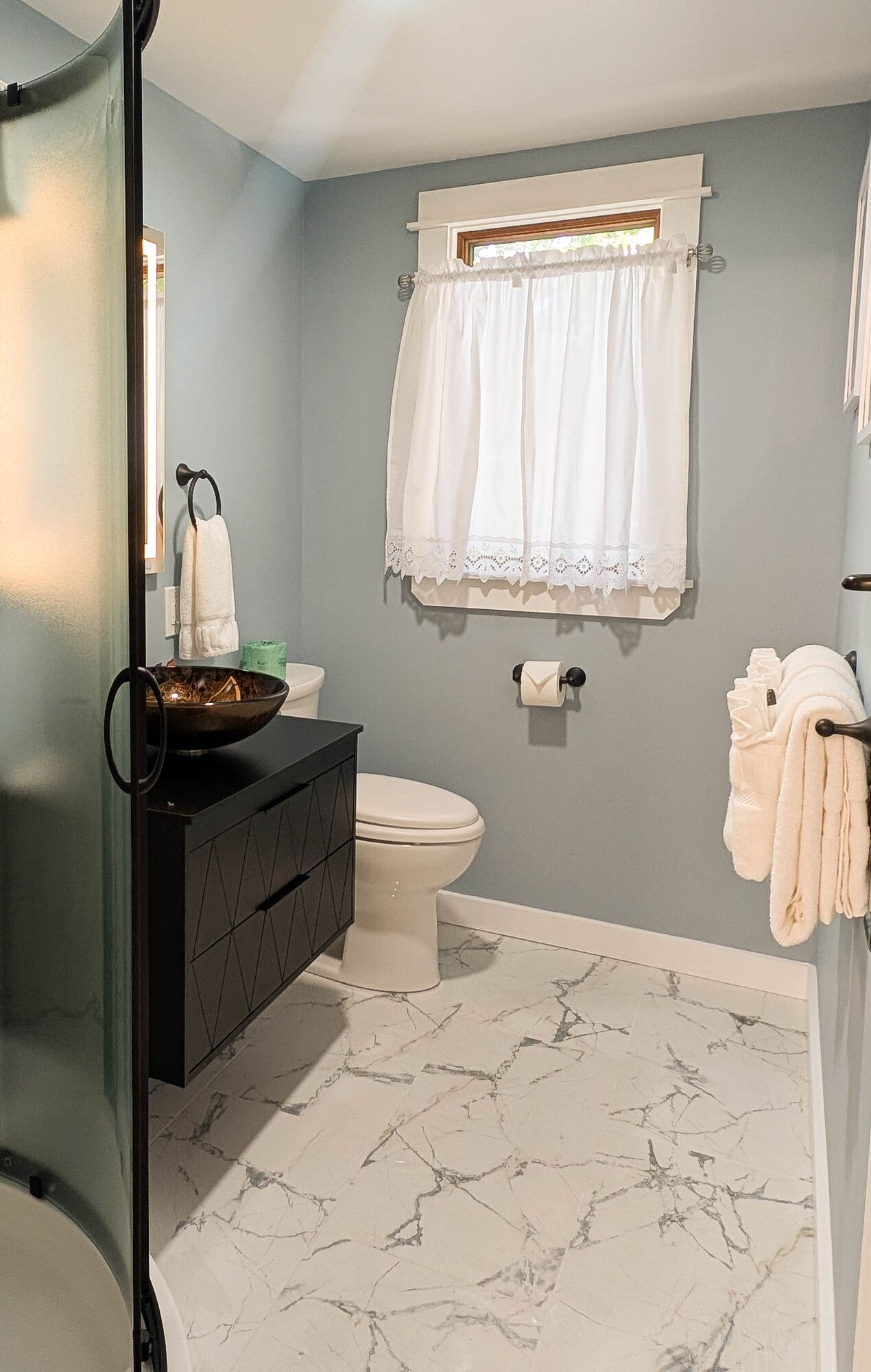 Bathroom with blue walls, white marble floor, black vanity, toilet, and a window with white curtains.