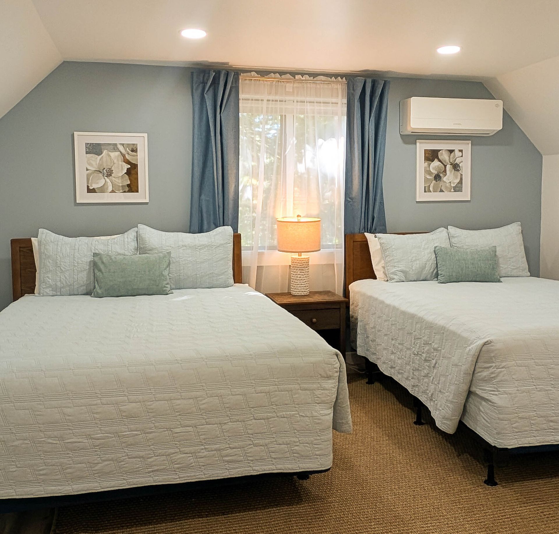 Two queen beds in a light blue bedroom with curtains, framed art, and an air conditioner.