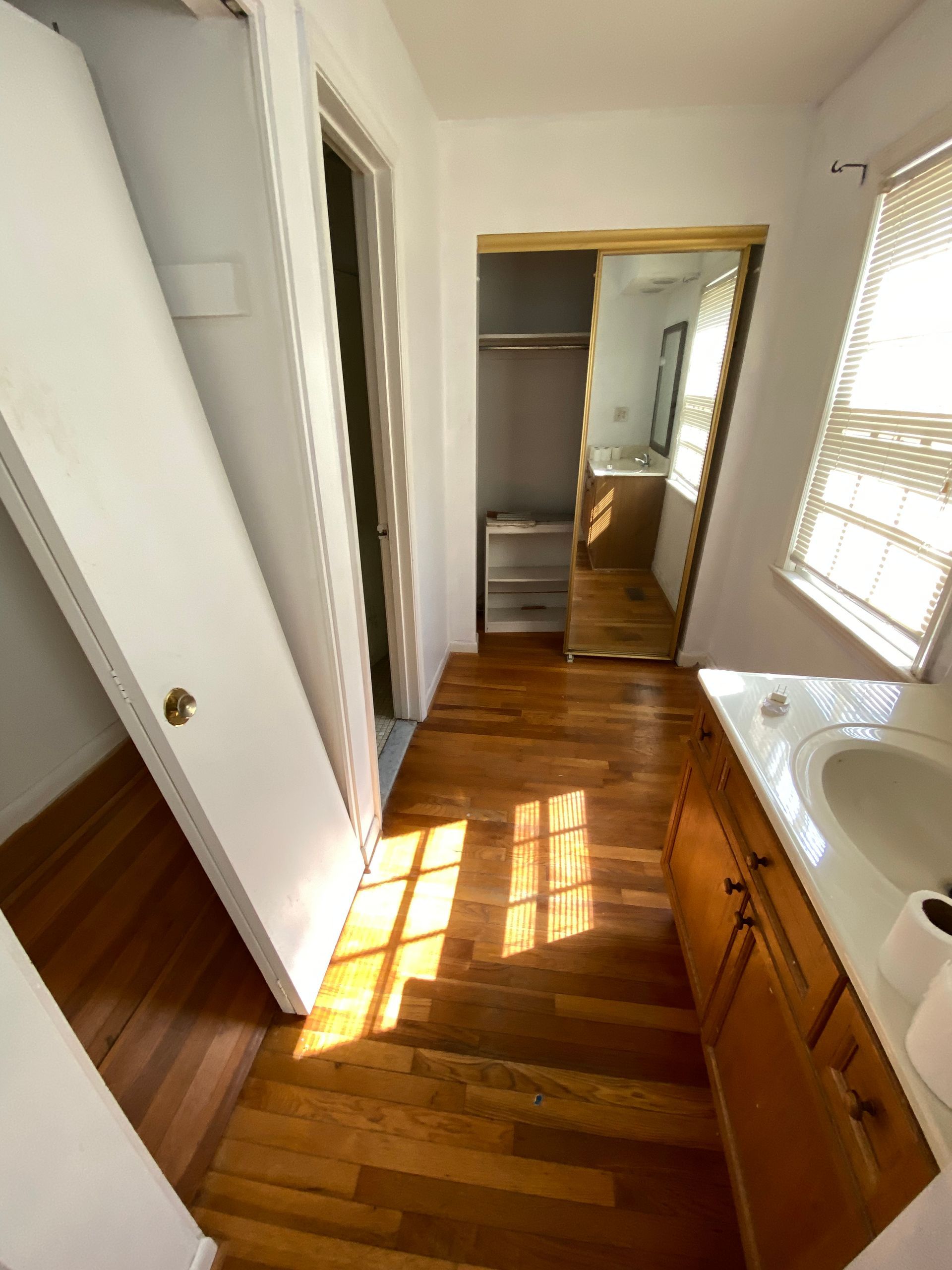 Bathroom with hardwood floor, wooden vanity, and closet. Sunlight streams through the window.