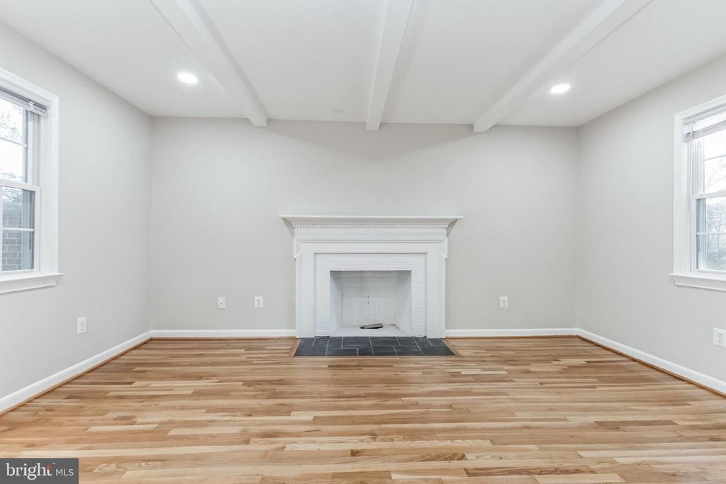 Empty living room with fireplace, hardwood floor, white walls, and exposed ceiling beams.