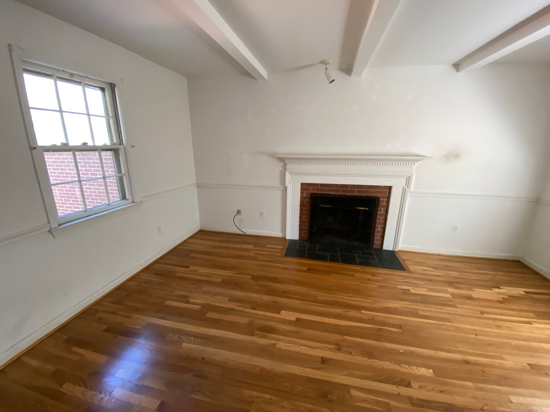 Empty room with wood floor, white walls, fireplace, and window with brick exterior visible.