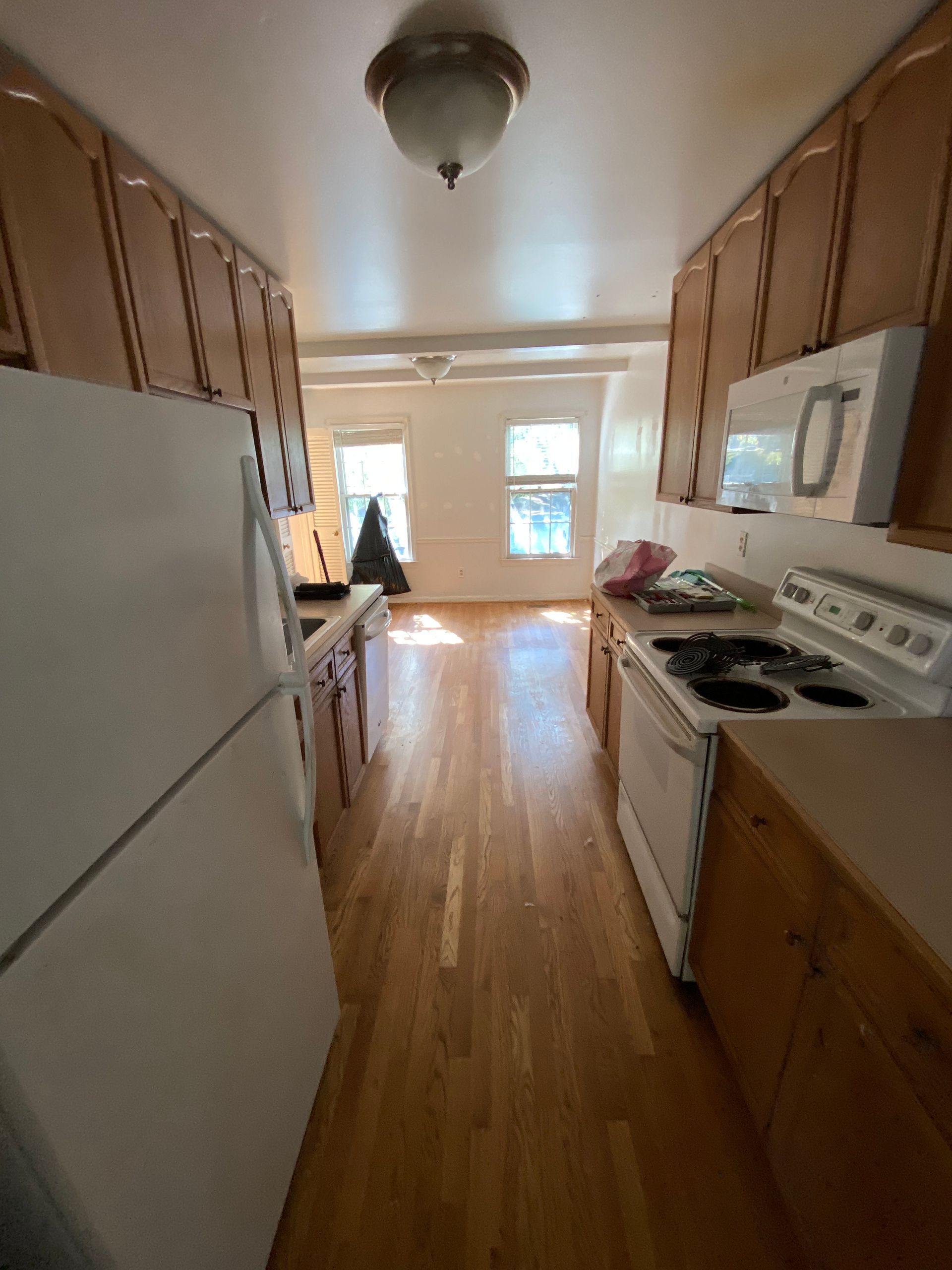 Narrow kitchen with wooden cabinets and floors, white appliances, leading to windows.