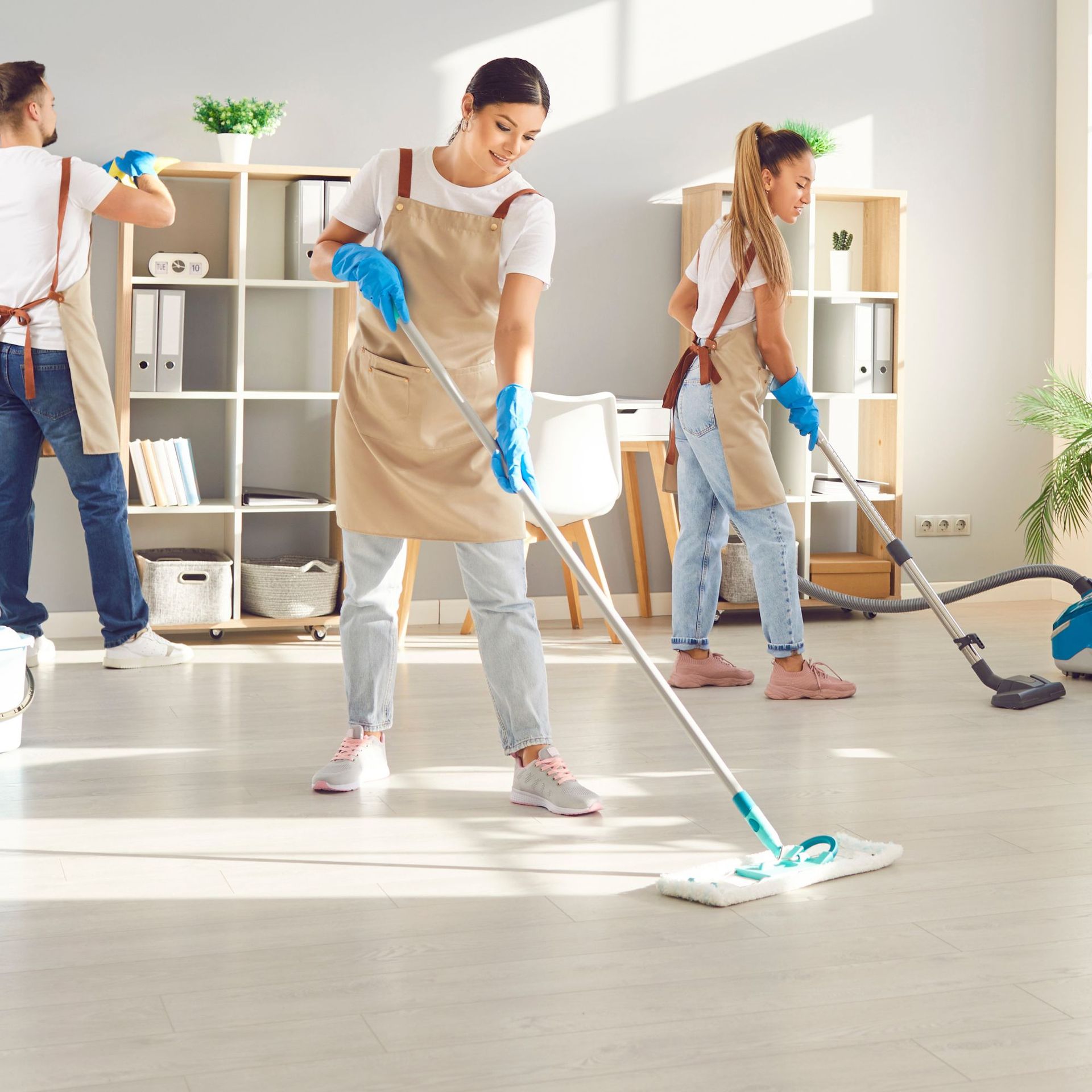 Three people cleaning an office: mopping, vacuuming, and dusting shelves.