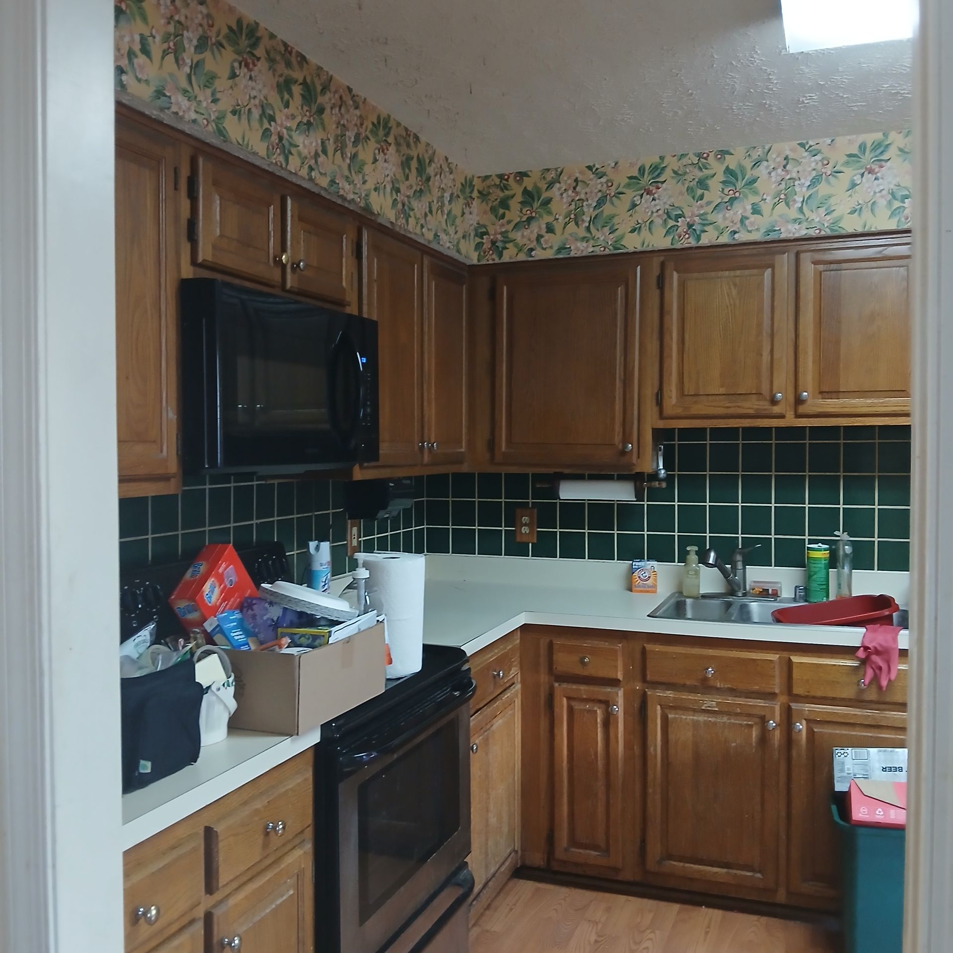 Kitchen with brown cabinets, microwave, stove, white counter, green tiled backsplash, and floral wallpaper.