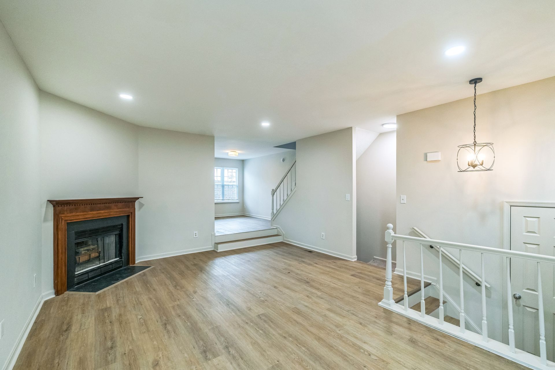 Empty living room with fireplace, stairs, and chandelier. Light wood floors, white walls.