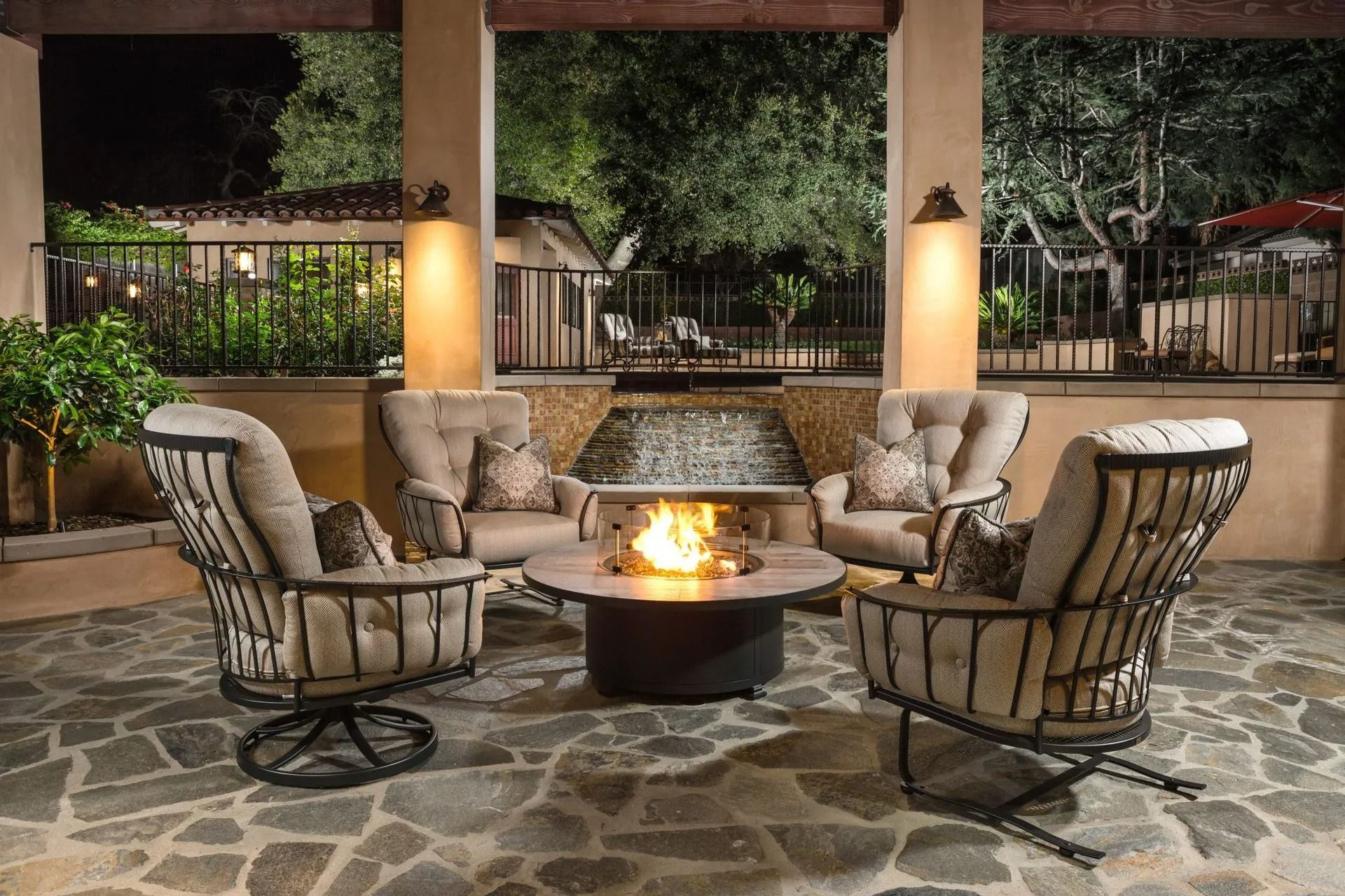 Outdoor seating area with fire pit and waterfall backdrop. Beige chairs, stone patio, nighttime.