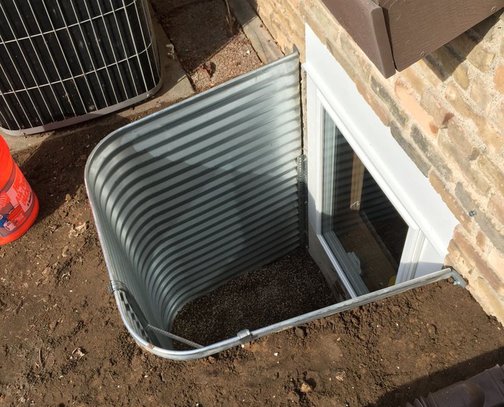 A window well is being installed on the side of a brick house.