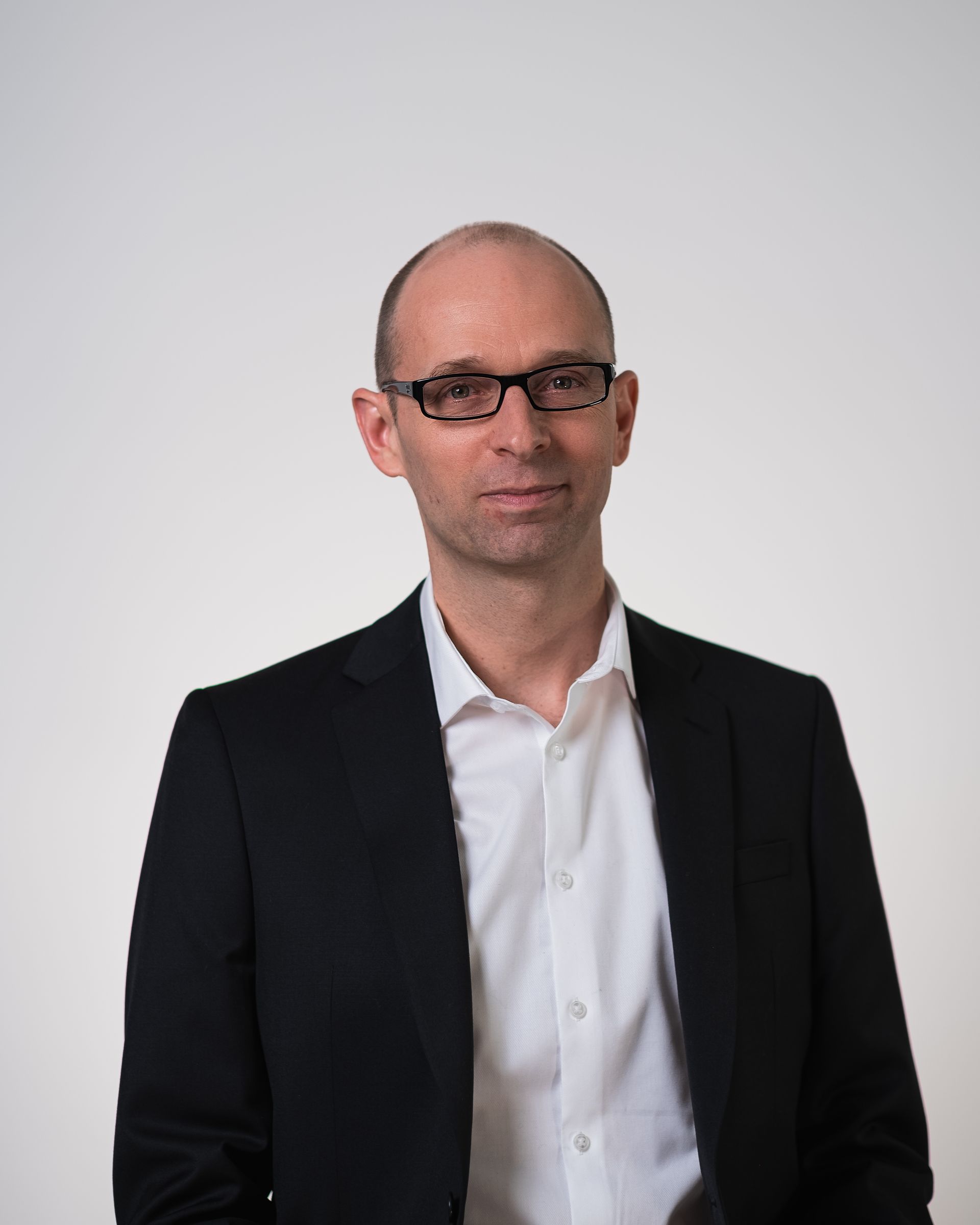 Un homme portant des lunettes et un costume se tient devant un mur blanc.