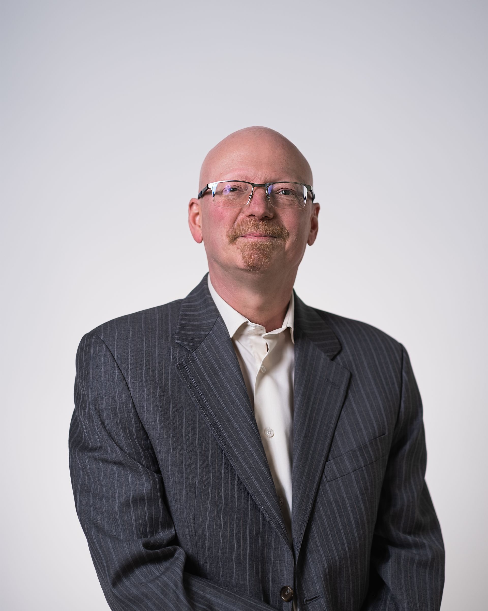 Un homme chauve portant des lunettes et un costume se tient devant un fond blanc.
