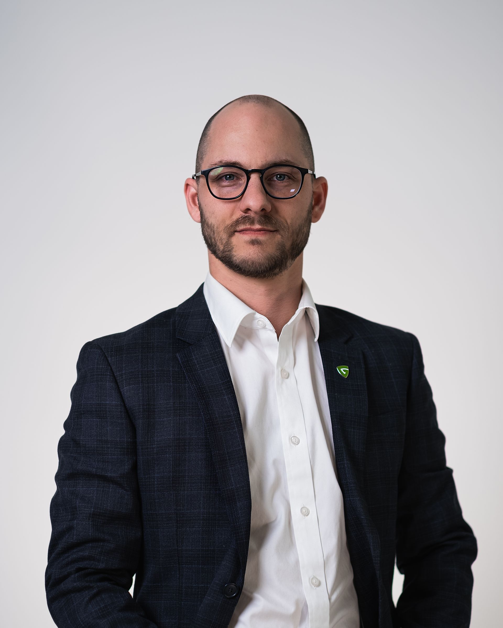 Un homme chauve portant des lunettes et un costume se tient devant un mur blanc.