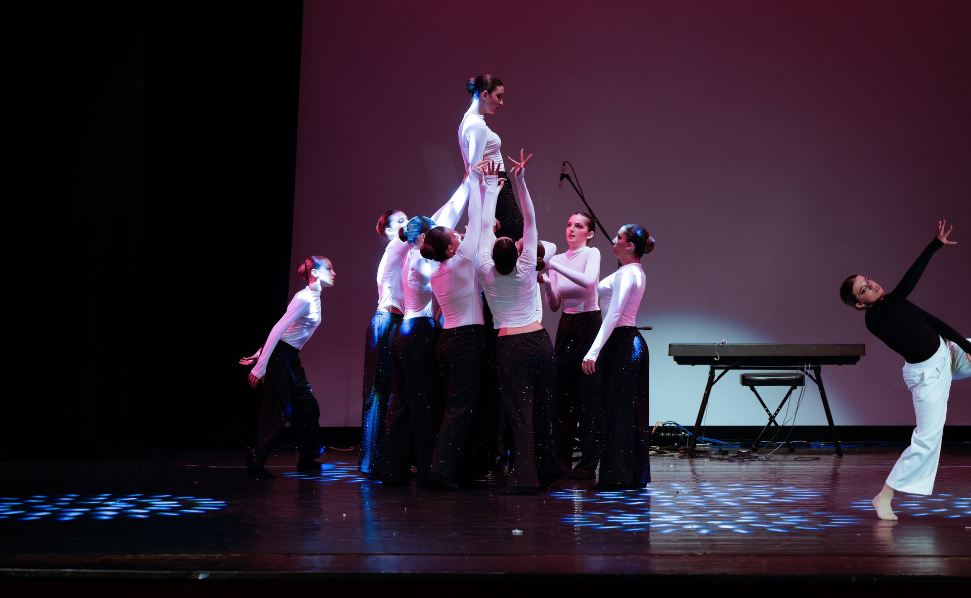 Ballerini che si esibiscono sul palco; uno in cima a una piramide umana, gli altri a fare da supporto. Pantaloni scuri, camicie bianche. Pianoforte in sottofondo.