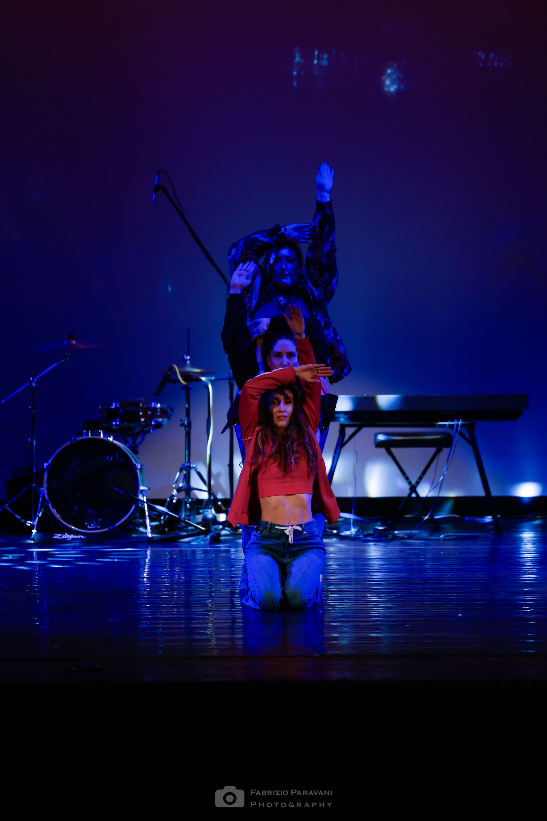 Tre persone sul palco, una in ginocchio e due in piedi, illuminate da una luce blu.