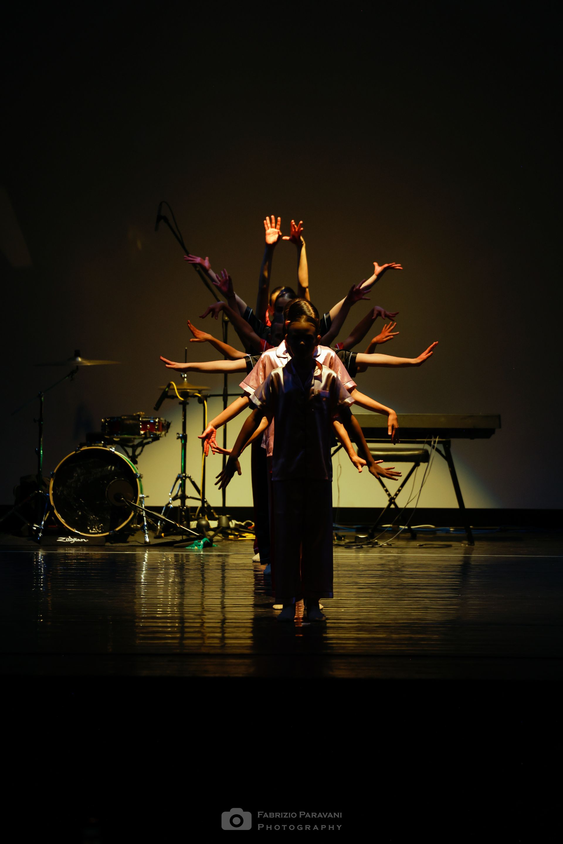 Ballerini sul palco con le braccia tese, creando l'illusione di molteplici braccia. Batteria e tastiera sullo sfondo.