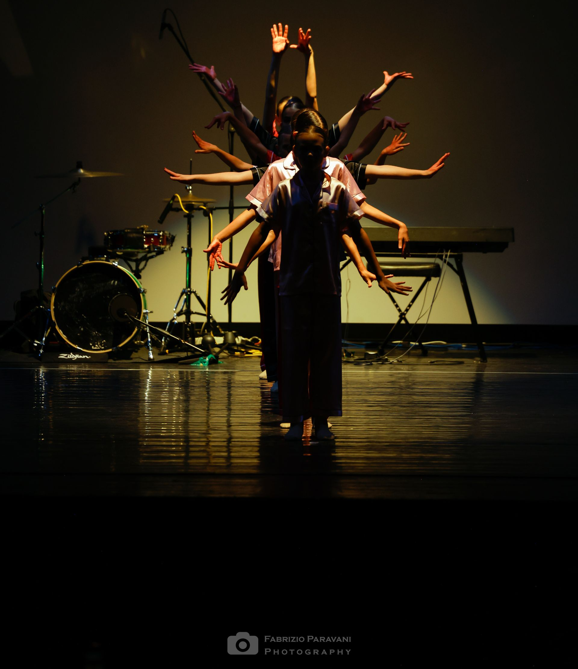 Ballerini con le braccia tese sul palco davanti a una batteria e una tastiera.