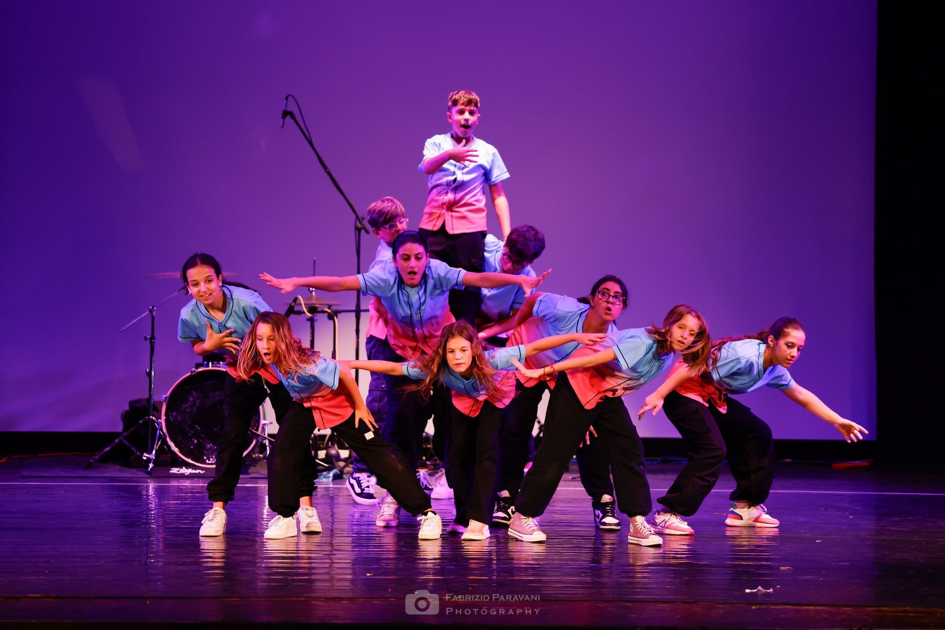 Gruppo di danza sul palco in formazione piramidale. Indossano camicie blu, pantaloni neri e scarpe da ginnastica. Sfondo viola.