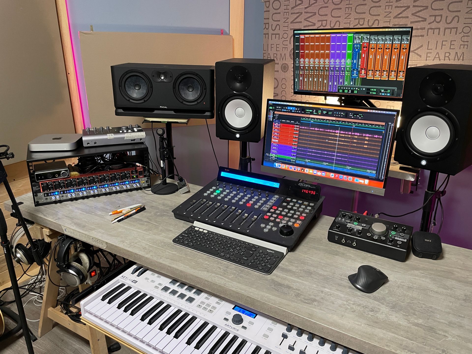 Studio di registrazione con monitor, mixer, tastiera e attrezzatura audio su una scrivania in legno.
