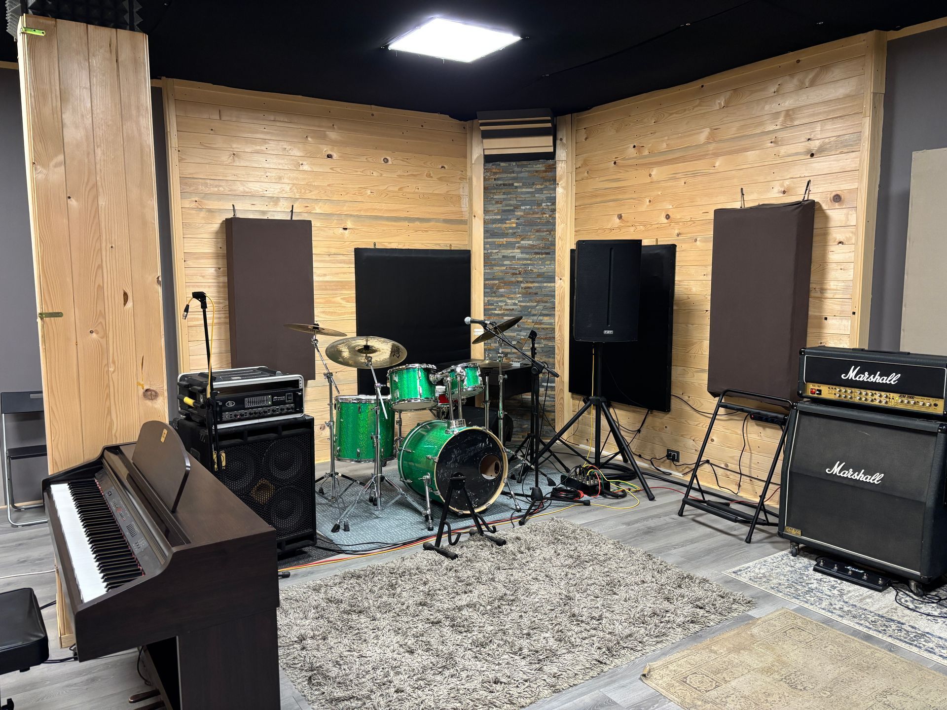 Studio musicale con batteria, pianoforte, amplificatori e pannelli acustici su pareti in legno.