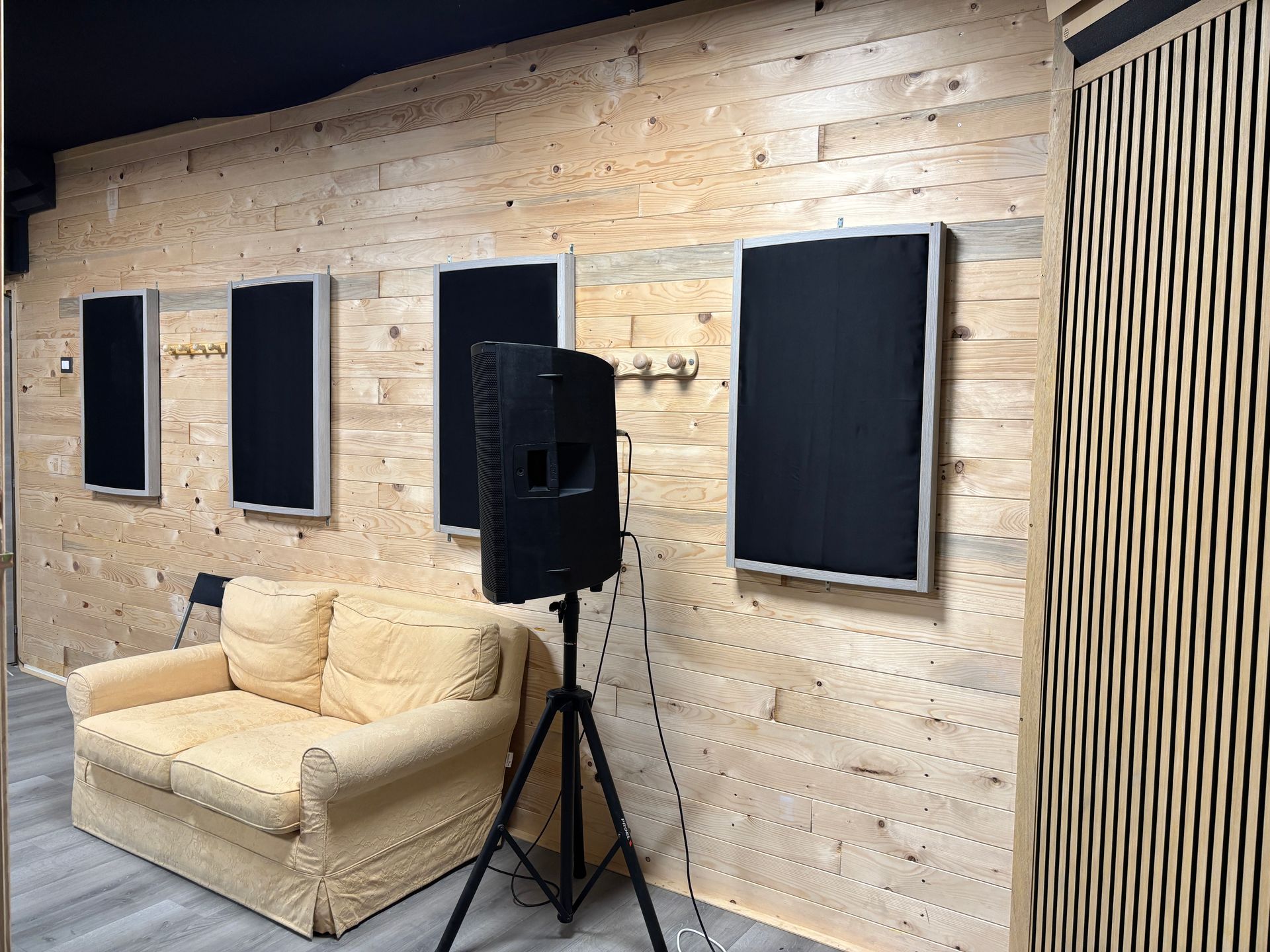 Interno di uno studio di registrazione con pannelli fonoassorbenti su una parete rivestita in legno, un altoparlante su un supporto e un divano.