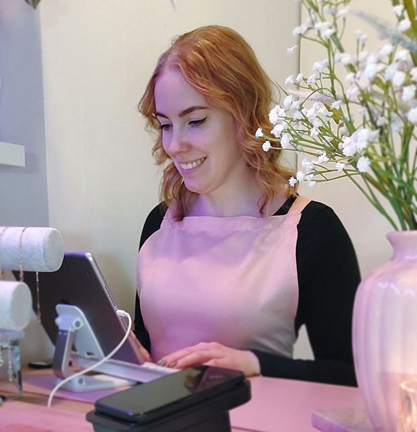 Rianne in een roze schort lacht terwijl ze een tablet gebruikt in een nagelstudio