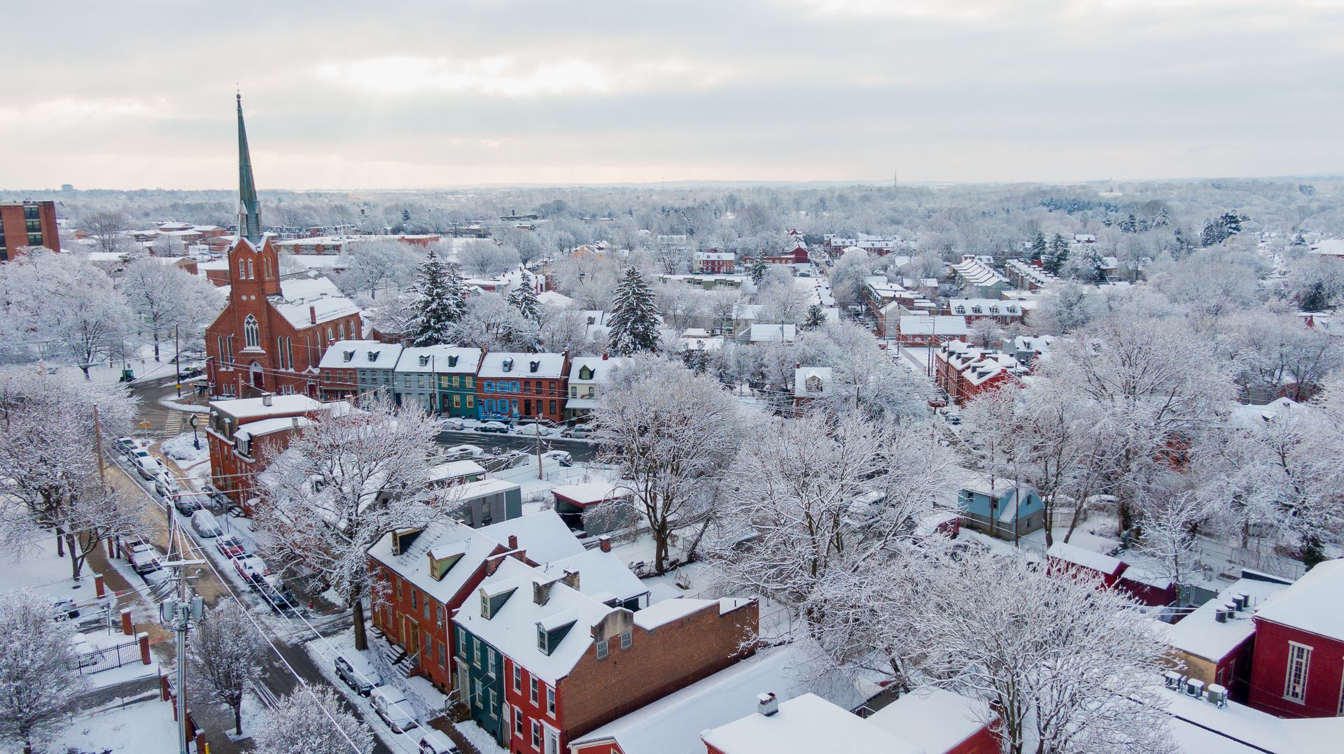 Snow-covered town; red brick buildings and church with a tall steeple. Trees are covered in snow under a cloudy sky.