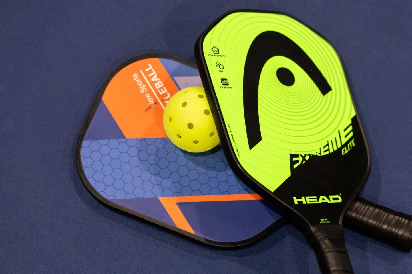 Two pickleball paddles, a yellow ball, blue surface. Green/black and blue/orange paddles.