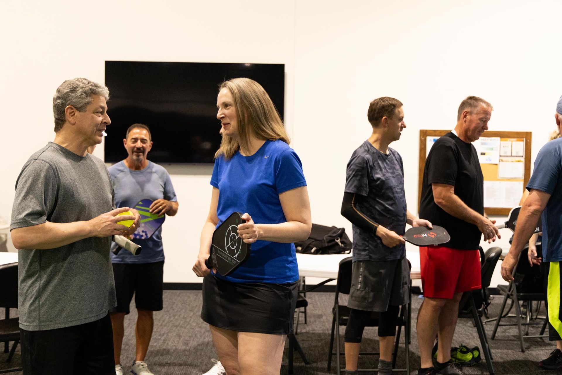People in a room, some holding pickleball paddles. Woman in blue shirt talks to man. Others stand nearby.