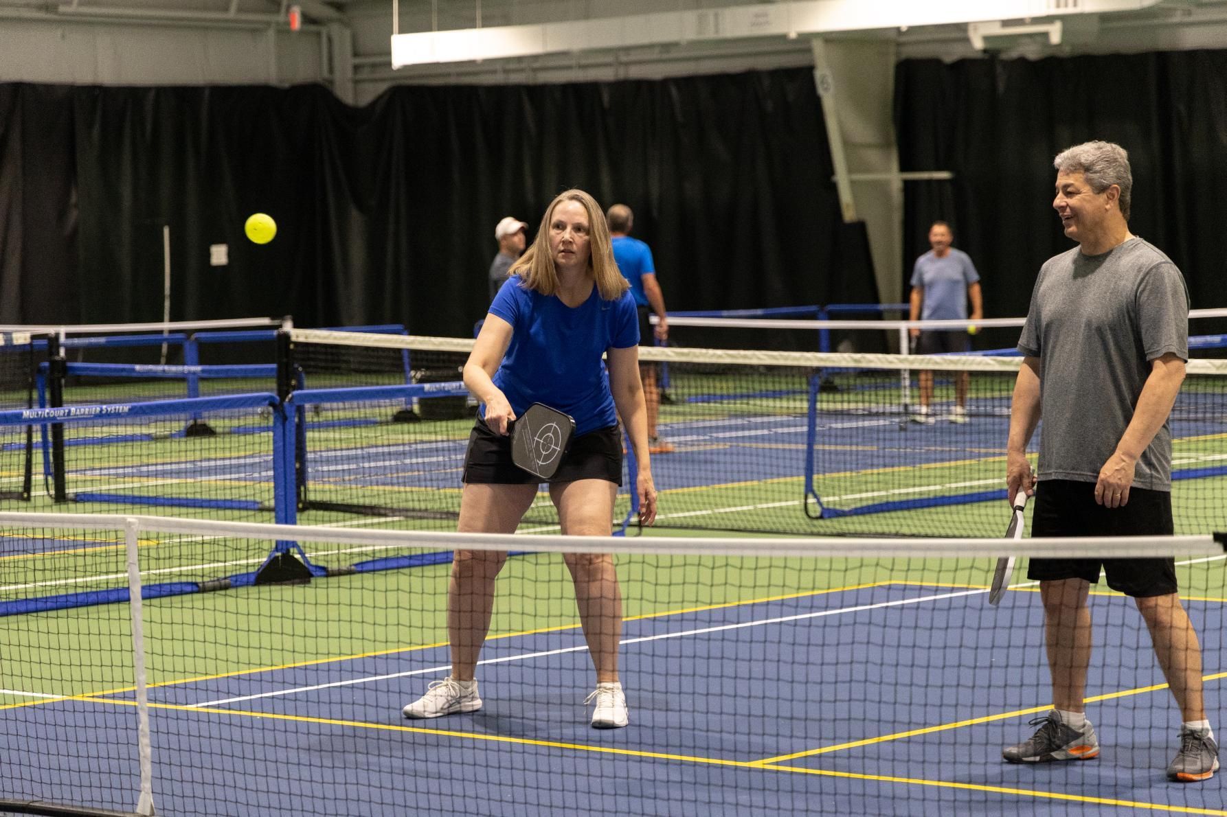 Two people playing pickleball on a blue court, a woman in blue hitting the yellow ball, man ready to return it.