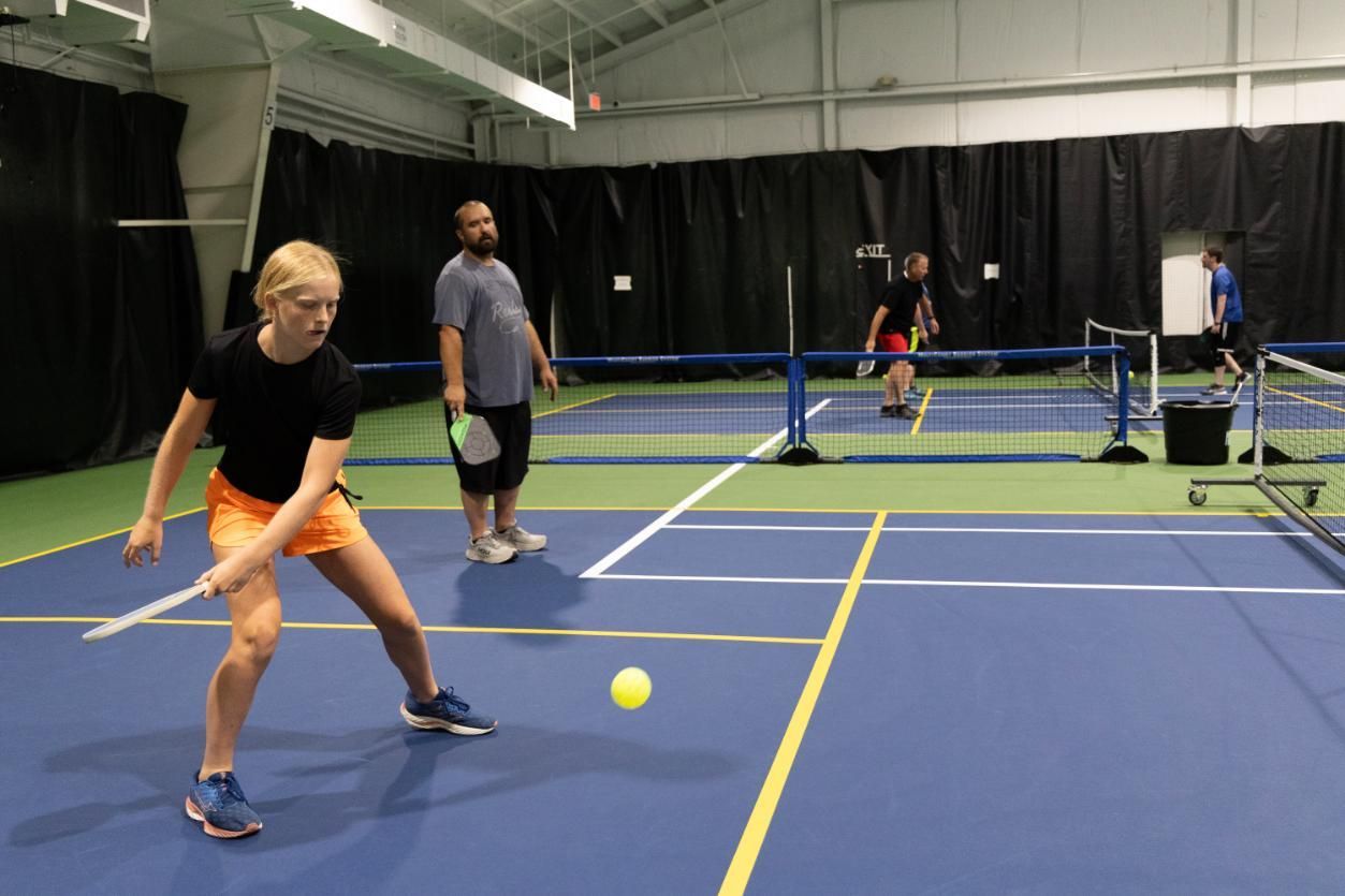 Woman in orange shorts playing pickleball; indoors on a blue court; others in background.