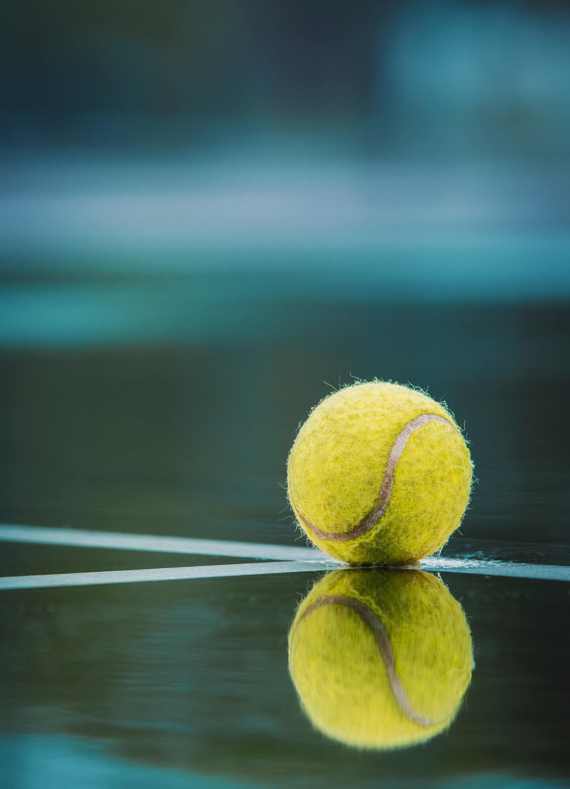 Tennis ball on a wet court, reflected in the surface. Yellow ball, white line, blue/green background.
