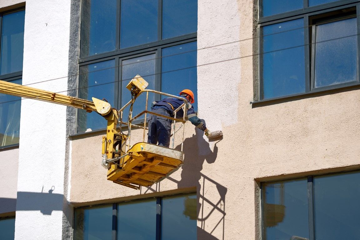 Worker on a suspended platform repairing a building exterior beside blue windows
