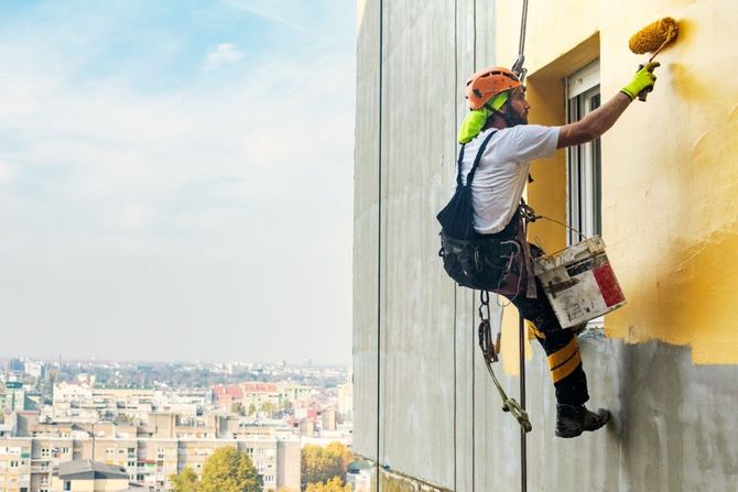 Rope-access worker painting a high-rise exterior wall with a roller, city skyline below