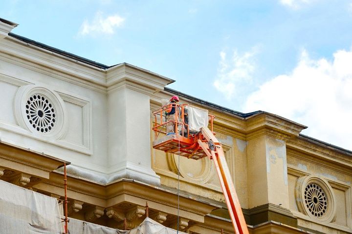 Worker on an orange lift repairing the ornate facade of a cream-colored building under a blue sky