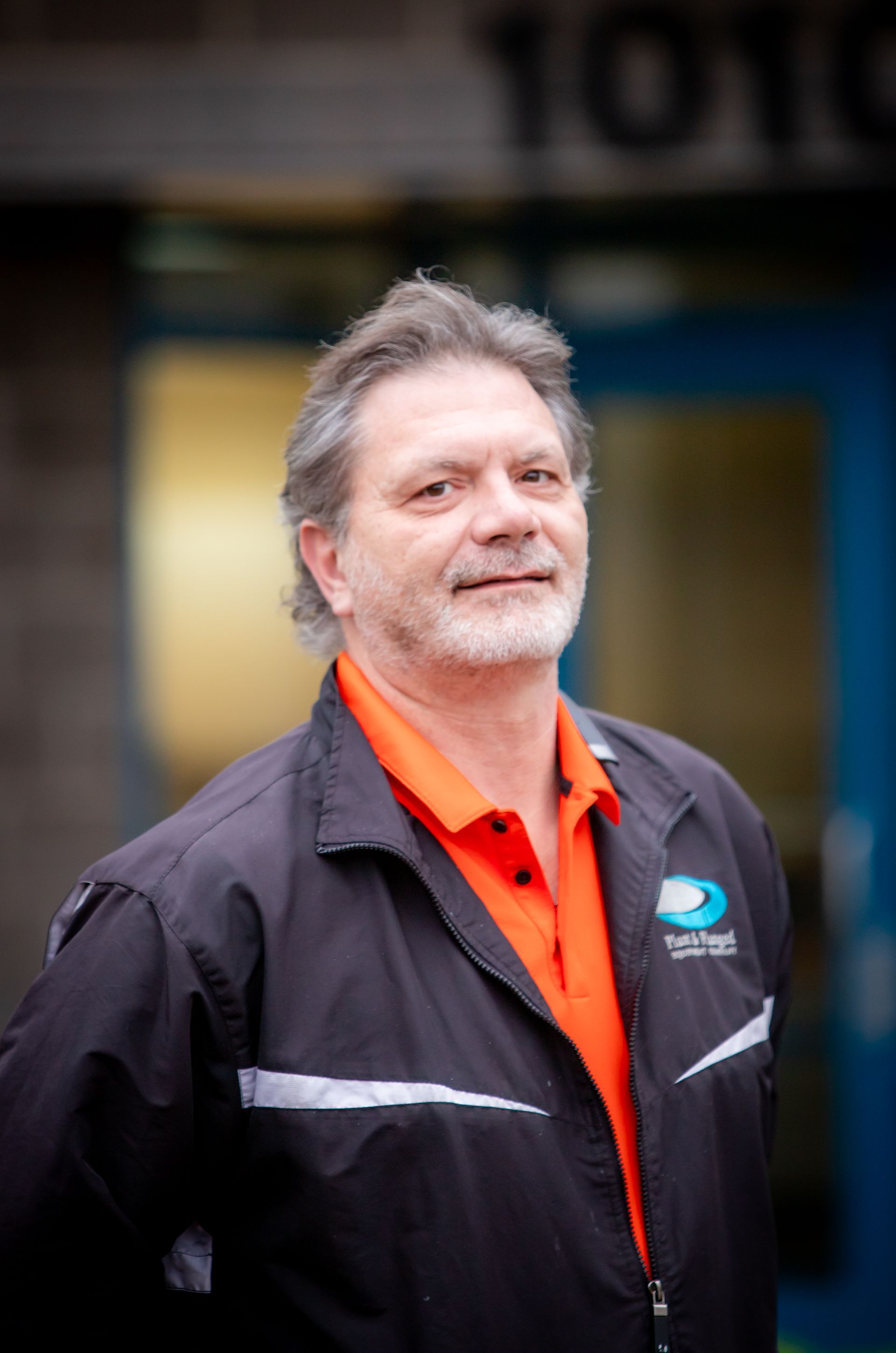 A man wearing an orange shirt and a black jacket stands in front of a building with the number 1010 on it