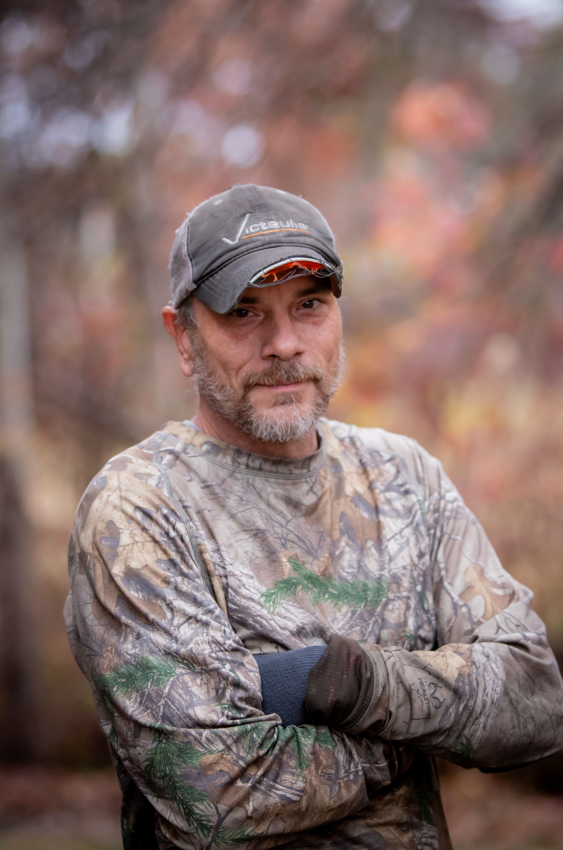 A man in a camouflage shirt and hat is standing with his arms crossed.