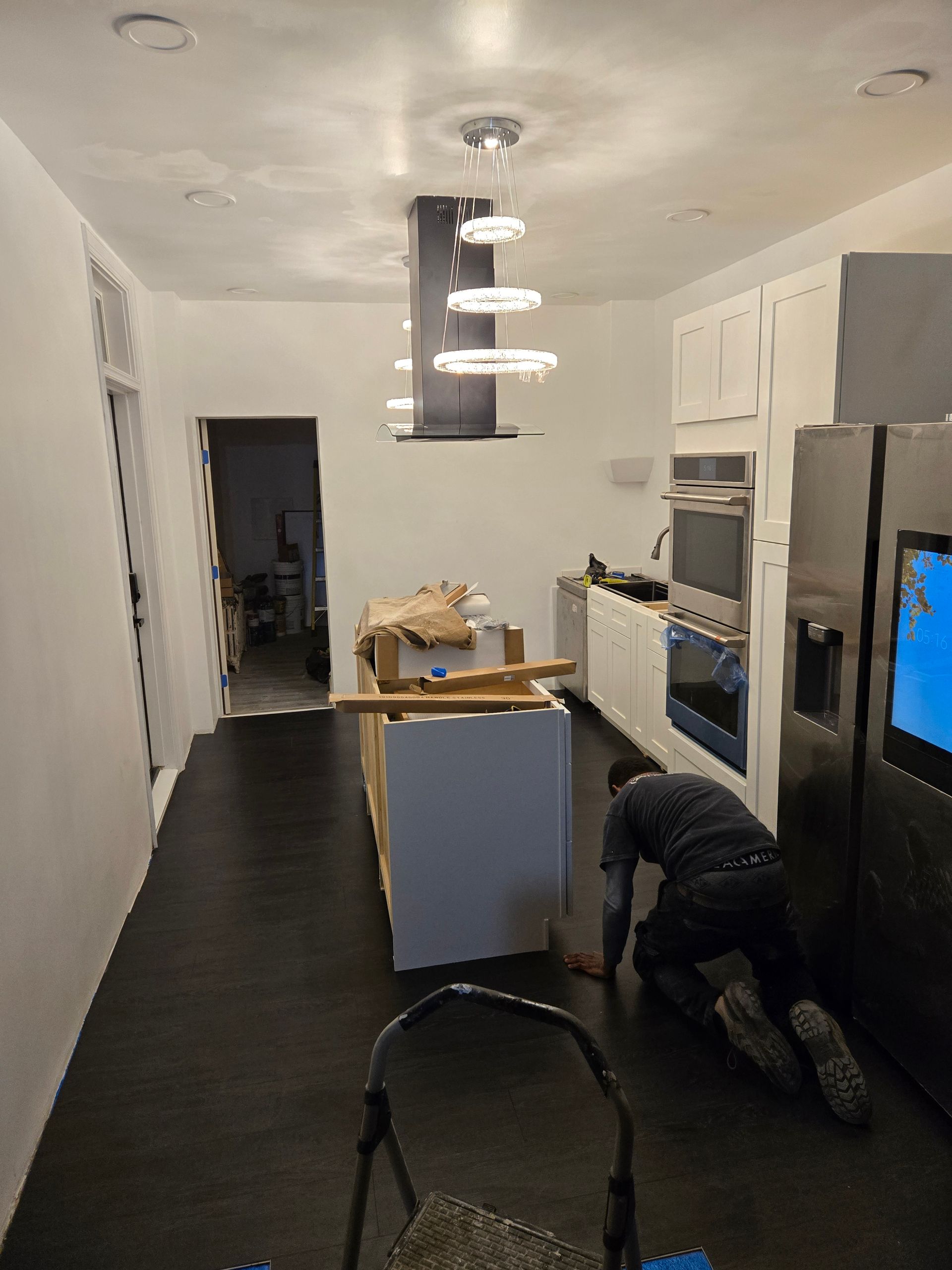Kitchen undergoing renovation, dark floor, white cabinets, island, worker kneeling, ladder, stainless steel appliances.