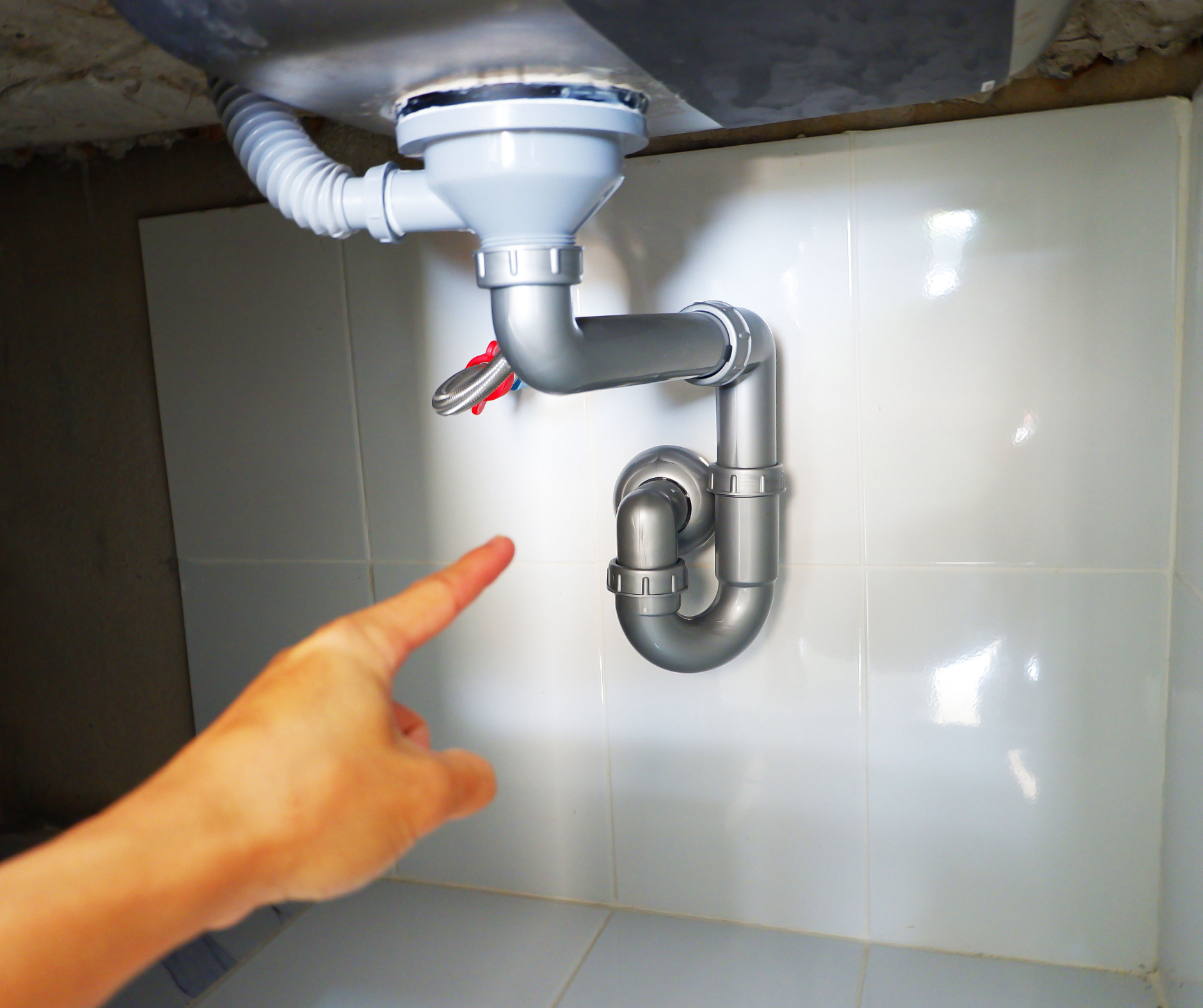 Hand pointing at a gray P-trap pipe under a white-tiled sink.