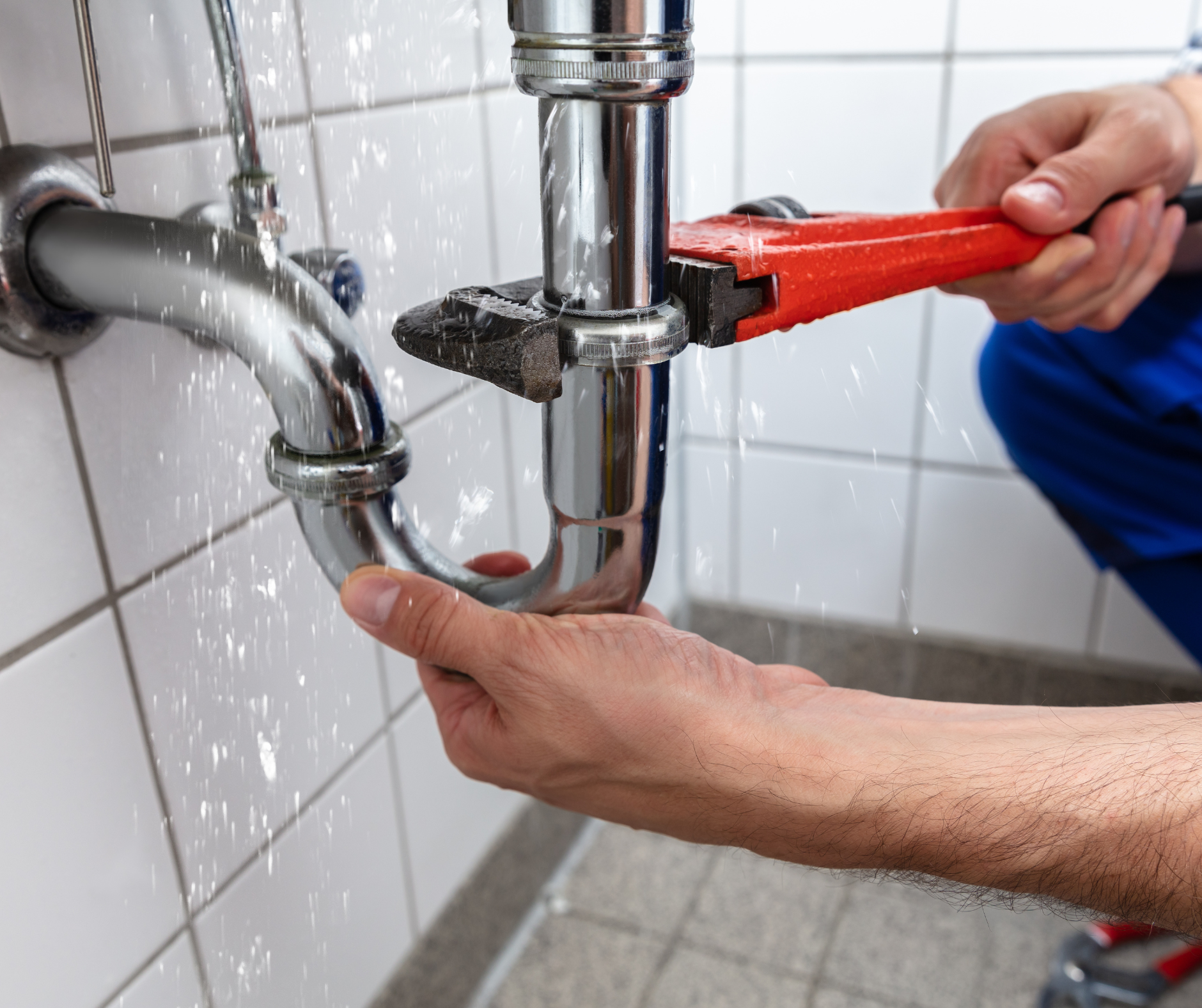 Plumber fixing a leaky chrome pipe with a wrench, water spraying, tiled wall.