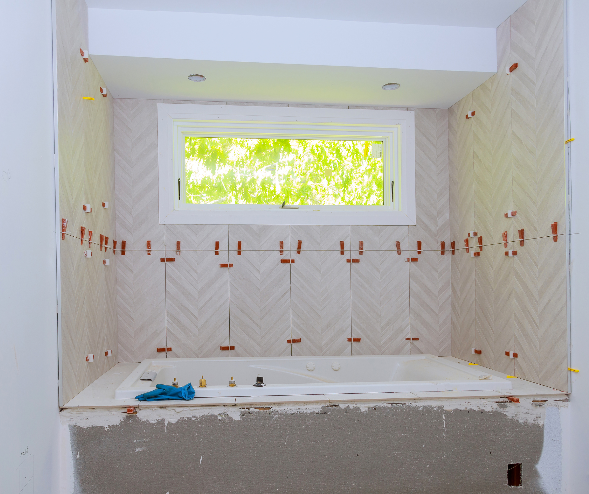 Bathroom under renovation, with white tiles installed around the tub, orange spacers, and a window.