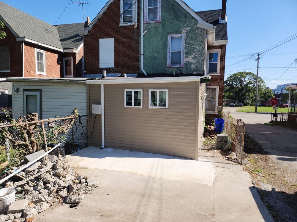 Back alley view: beige garage with two windows next to weathered brick and green painted buildings.