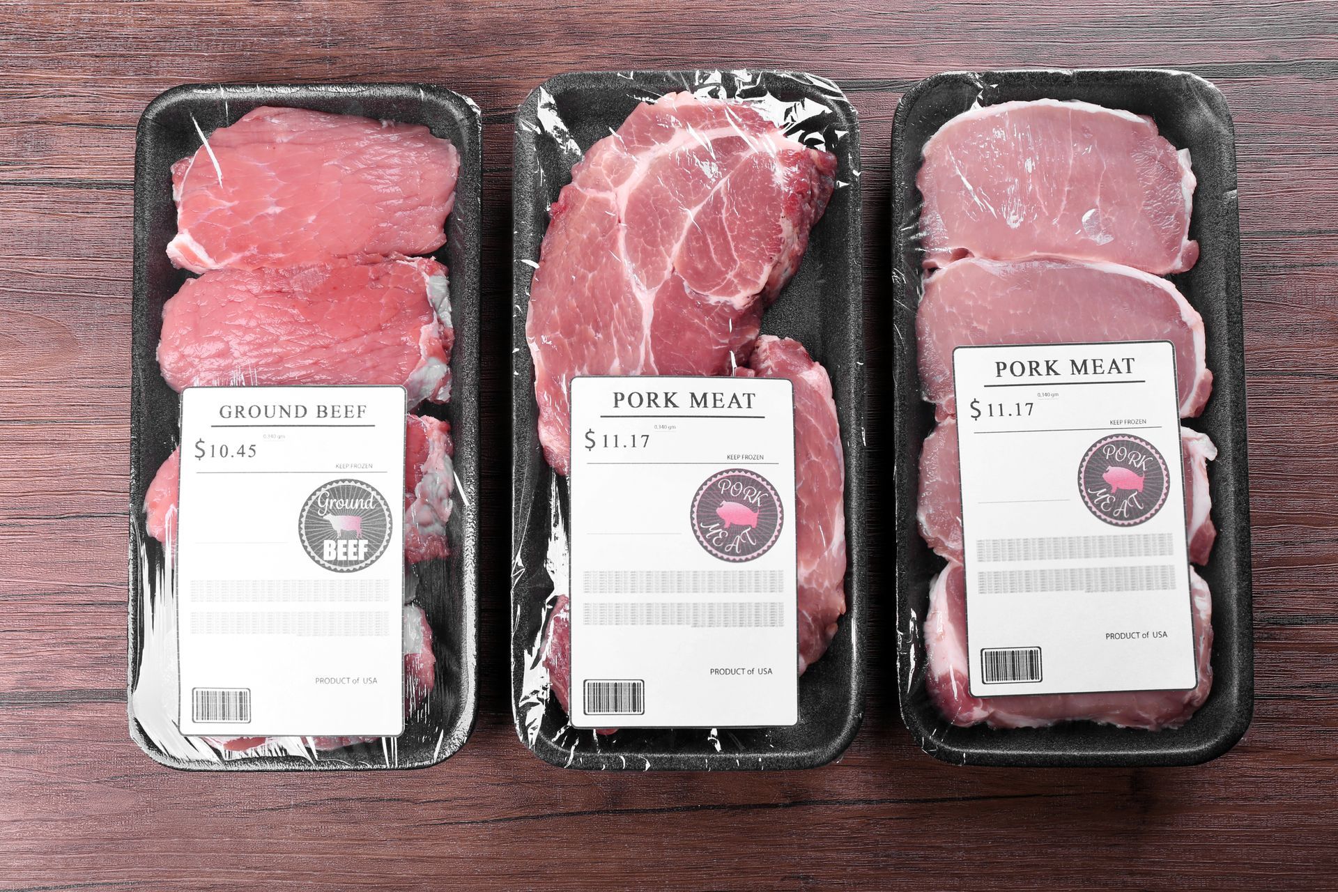 Three packaged raw beef steaks in black trays on a wooden table, each with a white label. Three packaged raw beef steaks in black trays on a wooden table, each with a white label.